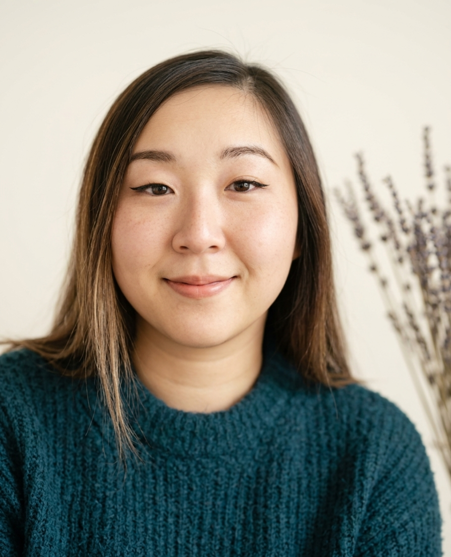 Woman with long brown hair wearing teal ribbed sweater, neutral beige background with dried lavender